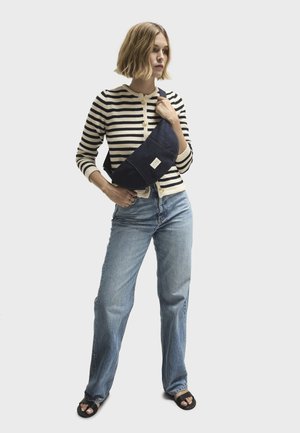 Borsa a tracolla in denim blu navy indossata sopra un cardigan a righe crema e nero. Il modello abbina jeans azzurri chiari e sandali neri. Design minimalista.