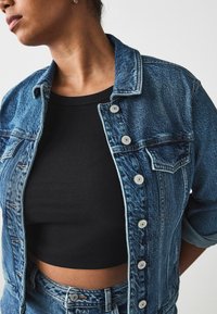 Denim jacket features a faded blue color, button front, and two chest pockets. Worn over a black ribbed crop top.