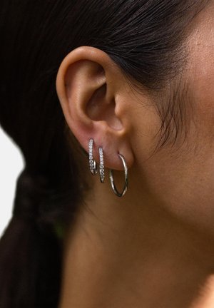 Nahaufnahme eines Frauenohrs mit drei silbernen Creolen, zwei kleineren Reifen mit eingearbeiteten Steinen und einer größeren schlichten Creole.