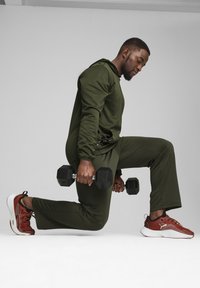 Tenue de sport verte avec un haut à capuche ajusté et un pantalon fuselé. Il tient un haltère noir, portant des baskets rouges et blanches avec un mesh.