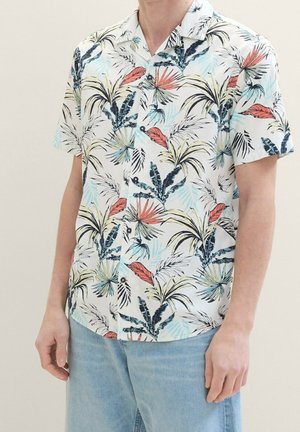 Homme portant une chemise à manches courtes avec un motif coloré de feuilles tropicales et un jean bleu clair.