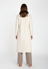 Donna con capelli castani di lunghezza spalle, che indossa un lungo cappotto crema, pantaloni beige a gamba larga e scarpe con tacco block beige, vista di spalle.