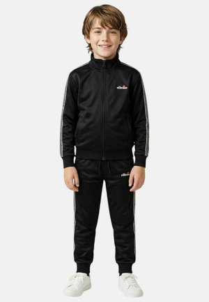 Garçon aux cheveux châtain clair portant un survêtement noir Ellesse et des baskets blanches, debout et souriant devant un fond uni.