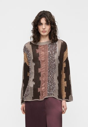 Kobieta o kręconych włosach, ubrana w wielokolorowy, teksturowany sweter i fioletowe spodnie, stojąca na tle jednolitego jasnego tła.