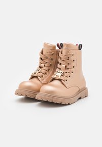 Stivaletti alla caviglia in ecopelle tan con lacci frontali, accessori in oro e linguetta a strisce. Suola in gomma resistente con battistrada texturizzato.