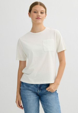 Mujer joven con cabello castaño claro que lleva una camiseta blanca de manga corta con un bolsillo texturizado y jeans azules, de pie frente a un fondo liso.