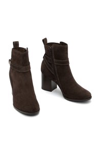 Botine din suede maro, cu închidere cu fermoar pe lateral, având o curea decorativă și un toc conic. Textură netedă și linii curate.