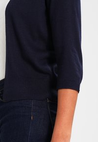 Marinblå cardigan tillverkad av mjukt stickat material, med ribbad fåll och trekvartslånga ärmar, kombinerad med en ljusfärgad figurnära topp.