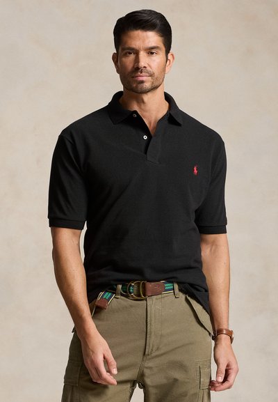 Mann trägt schwarzes Poloshirt mit rotem Logo, grüner gestreifter Gürtel, khakifarbene Cargo-Hose und braune Armbanduhr vor beigem Hintergrund.