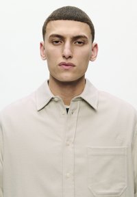 Young man with short brown hair wearing a beige button-up shirt with a chest pocket and embroidered "W" on a plain white background.