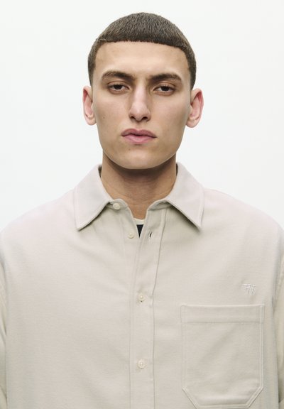 Jeune homme avec des cheveux bruns courts portant une chemise beige à boutons avec une poche poitrine et un petit "W" brodé sur un fond uni.