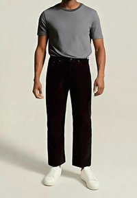 Homme portant une chemise grise à manches courtes, un pantalon noir et des baskets blanches à enfiler, debout devant un fond clair uni.