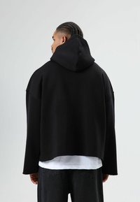 Zwarte oversized hoodie met capuchon met trekkoord, onafgewerkte zomen en verlaagde schouders. Gedragen over een wit gelaagd shirt, gezien vanaf de achterkant.