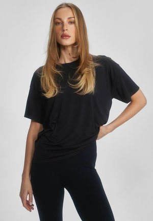 Kobieta o długich blond włosach, ubrana w luźną czarną koszulkę i czarne spodnie, stojąca z jedną ręką na biodrze na jednolitym tle.