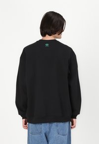 Sweatshirt preto com punhos e barra canelados, apresentando um pequeno logotipo verde da Adidas na parte de trás do pescoço, usado com jeans azul claro.