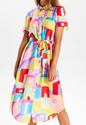 Femme portant une robe-chemise colorée boutonnée avec manches courtes et ceinture, avec des maisons abstraites aux tons rouge, bleu, violet, jaune et vert.