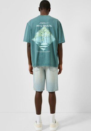 Mężczyzna o ciemnej karnacji, ubrany w turkusowy t-shirt z grafiką palmy i napisem „Into Brazil”, jasne jeansowe szorty, białe skarpetki i białe trampki, stojący tyłem.