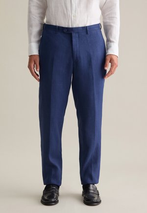 Pantalones de lino azul marino con corte recto, con frente plano y dos botones. Llevados con una camisa de manga larga blanca y zapatos negros.