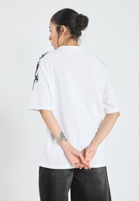 Camiseta blanca de manga corta con un corte relajado y un diseño de llamas negras en los hombros. Textura suave, con la parte de atrás lisa, combinada con jeans oscuros.