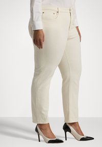 Krémové džíny s úzkým střihem, vybavené zipem, viditelným švem a hladkou strukturou, doplněné černobílými lodičkami.
