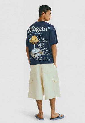 Ung mand står med hænderne i lommerne iført en marineblå t-shirt med affogato-grafik og beige vidde shorts med lommer og blå flipflops.