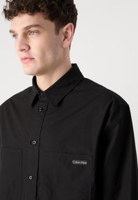 Juodas mygtukų užsegamas marškinys iš medvilnės mišinio, su išplatintu apykakle, dviem priekinėmis kišenėmis ir maža balta Calvin Klein logo kairėje kišenėje.