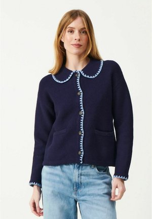 Kvinde iført en marineblå cardigan med knapper og lys blå bølget kant, kombineret med lyseblå jeans, stående foran en ensfarvet baggrund.