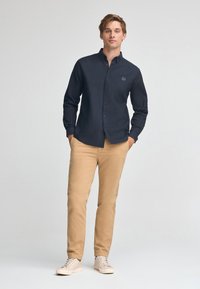 Camicia button-up a maniche lunghe di un blu scuro, con un piccolo logo, abbinata a pantaloni chinos beige e sneakers chiare. Tessuto morbido, vestibilità classica.