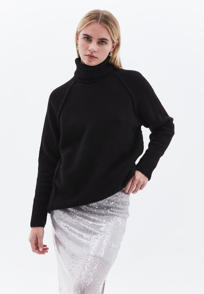 Zwarte coltrui van gestructureerde breigoed, met geribde manchetten en zoom. Draagt over een glanzende zilveren paillettenrok.