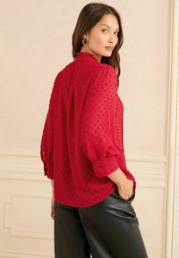 Blusa rossa con maniche a sbuffo e motivo a puntini, scollatura rotonda e una vestibilità leggermente morbida, abbinata a pantaloni neri sartoriali.