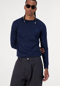 Navy long-sleeve shirt with a blue logo on the chest, paired with dark gray baggy pants featuring side pockets and a snap button closure.