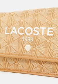 Portafoglio Lacoste in materiale beige testurizzato con motivi geometrici, dotato di un logo bianco prominente e una chiusura a bottone color crema.