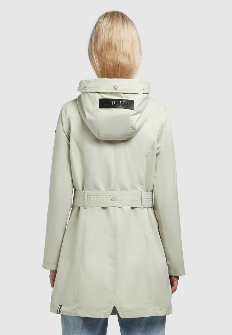 Giacca impermeabile verde chiaro con cappuccio, vita cinturata, ventola posteriore e un logo nero sulla scollatura. Texture liscia e design elegante.