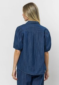 Tamsiai mėlynas džinsinis marškinis, su pūkuotomis trumpomis rankovėmis, klasikinio kirpimo apykakle ir išlenktu hemu. Minkšta tekstūra su kasdiene išvaizda.