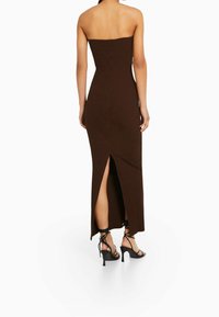 Robe maxi marron, sans bretelles, en maille côtelée avec une fente haute à l'arrière. Le mannequin porte des escarpins à lacets noirs, mettant en valeur une silhouette ajustée.