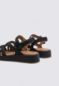 Sandalias de plataforma trenzadas negras con un diseño de tiras texturizadas, hebilla ajustable y plantilla acolchada marrón. Base de plataforma completamente negra.