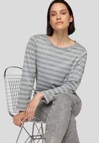 Graues Langarmshirt mit horizontalen Streifen, weichem Textil, rundem Halsausschnitt und lockerem Schnitt. Getragen mit grauen Jeans, sitzend auf einem Drahtstuhl.