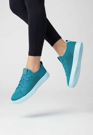 Persoon draagt turquoise gebreide sneakers met lichtblauwe zolen en zwarte leggings, vastgelegd in de lucht tegen een effen grijze achtergrond.