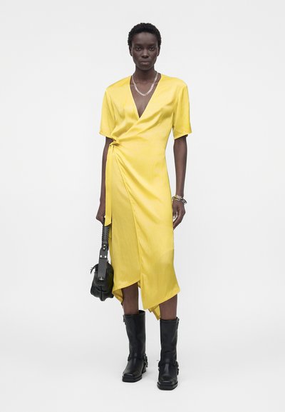 Γυναίκα που φοράει κίτρινο φόρεμα τύπου wrap με κοντά μανίκια, μαύρα μπότες μέχρι τη μέση της γάμπας, ασημένιο κολιέ, βραχιόλια και κρατά μια μαύρη τσάντα.