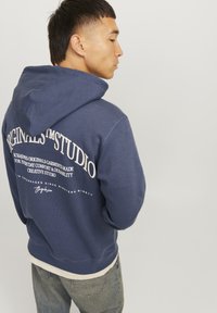 Sudadera con capucha de color azul marino con texto blanco en la parte posterior, puños y dobladillo acanalados. Detalles de logotipo y marca visibles.