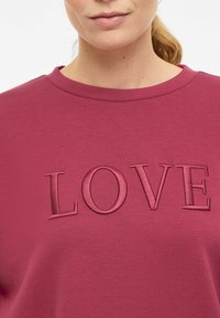 Sweat-shirt bordeaux avec col rond et motif "LOVE" embossé de la même couleur. Tissu doux avec une texture lisse.