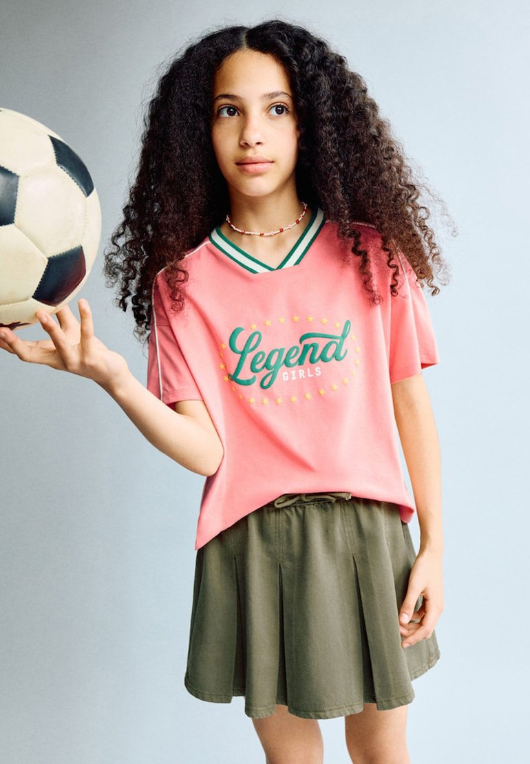 Fille aux cheveux bouclés portant un t-shirt rose « Legend Girls » et une jupe olive, tenant un ballon de football noir et blanc d'une main.