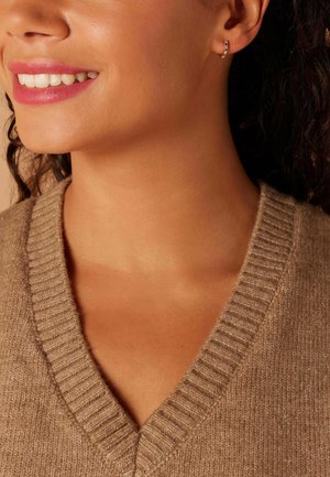 Beige gebreide trui met een V-hals, voorzien van ribbeldetails. Geaccentueerd door kleine hoepeloorbellen met gekleurde edelstenen. Glad huidtint.