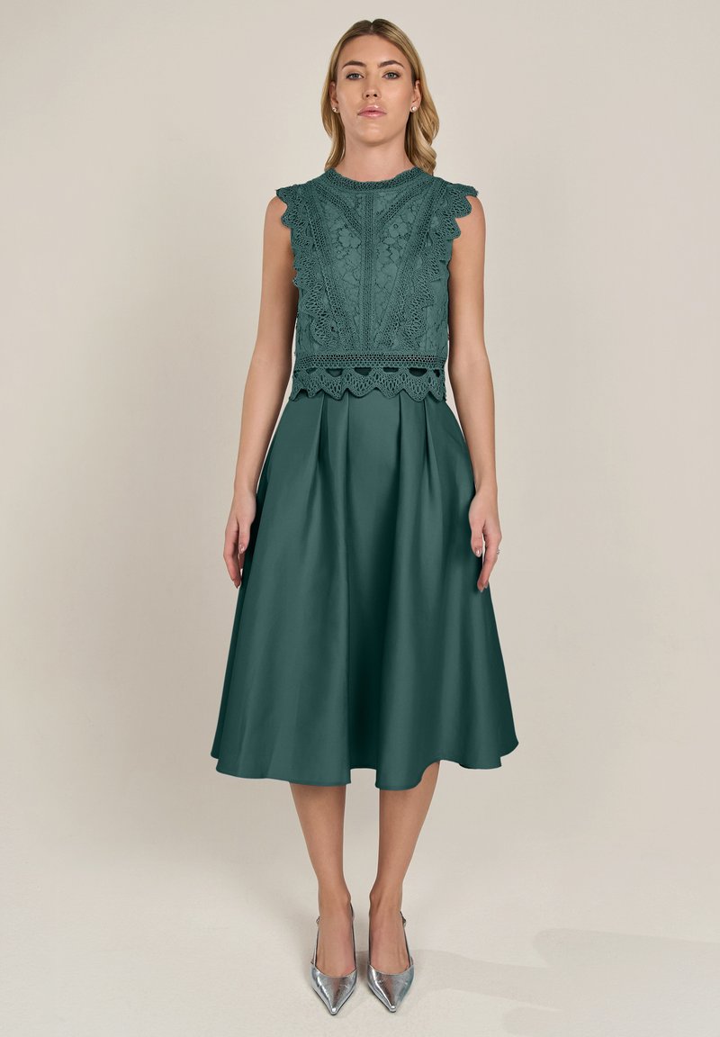 Teealfarbenes Kleid mit Spitzen-Oberteil, gewelltem Saum und plissiertem Rock. Getragen mit silbernen spitzen High Heels. Glatte Stofftextur.