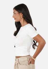 Witte t-shirt met korte mouwen en een aangesloten ontwerp, voorzien van een klein rood logo aan de linkerkant, gecombineerd met beige hoge taille broek.