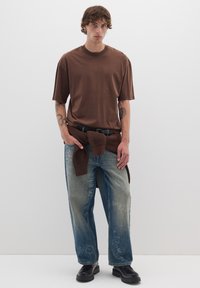 T-shirt in cotone marrone con maniche corte, abbinato a jeans in denim chiaro strappati con testo ricamato, e scarpe nere chunky.