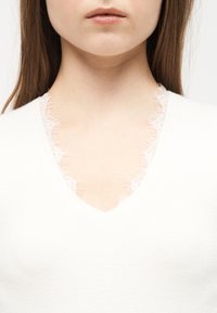 Κοντινό πλάνο στον λαιμό και το κάτω μέρος του προσώπου μιας γυναίκας που φοράει λευκό τοπ με V-λαιμόκοψη και λεπτή δαντέλα κατά μήκος της γραμμής του λαιμού.