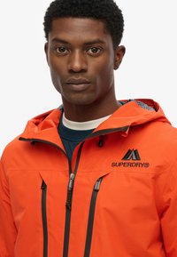 Jasnopomarańczowa wodoodporna kurtka z kapturem, z czarnymi zamkami błyskawicznymi i logo. Posiada dwie przednie kieszenie oraz wzorzystą podszewkę.