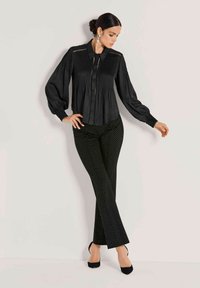 Zwarte geplooide blouse met een kraag en knoopsluiting aan de voorkant, gecombineerd met strakke zwarte broek met witte stippen en hakken.