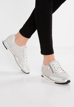 Feet wearing silver metallic lace-up sneakers with side zipper and perforated details, paired with black fitted pants.